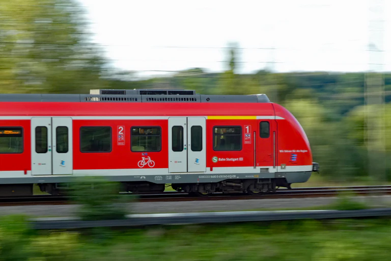 Ein roter S-Bahn-Zug vor einer bewaldeten Landschaft, die durch Bewegungsunschärfe verschwommen ist. — © Symbolbild CatalpaSpirit (CC BY-SA 4.0)